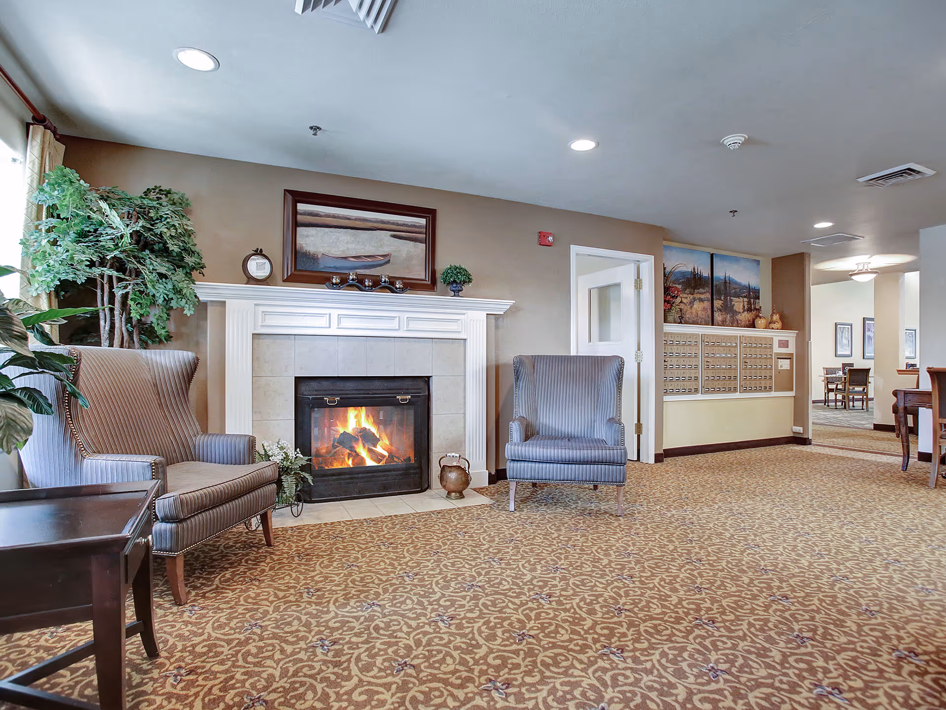 A cozy senior living common room with a lit fireplace, two upholstered armchairs, plants, and a wall of mailboxes.