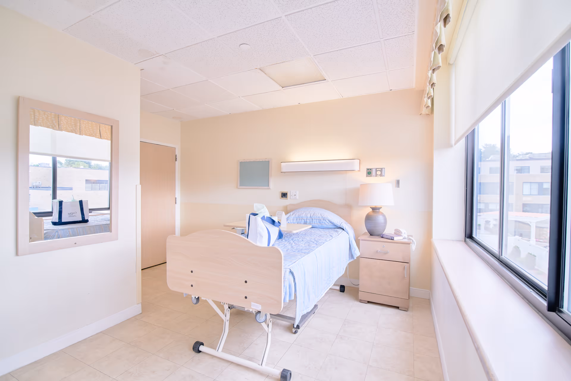 A bright and clean senior living facility bedroom with a hospital-style bed covered in light blue bedding, a bedside table with a lamp and phone, a large window letting in natural light, a mirror on the wall, and a door in the background.