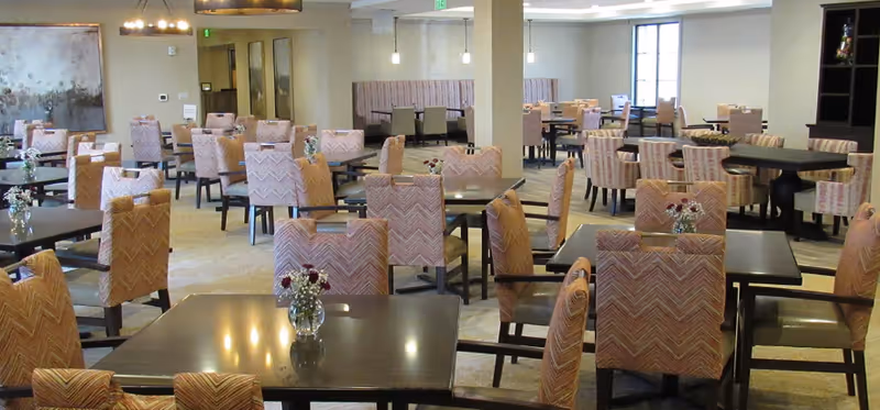 A spacious dining room with multiple tables and cushioned chairs arranged neatly. Each table has a small vase with flowers. The room is well-lit with ceiling lights and has a neutral color scheme with beige walls and carpeted floors.