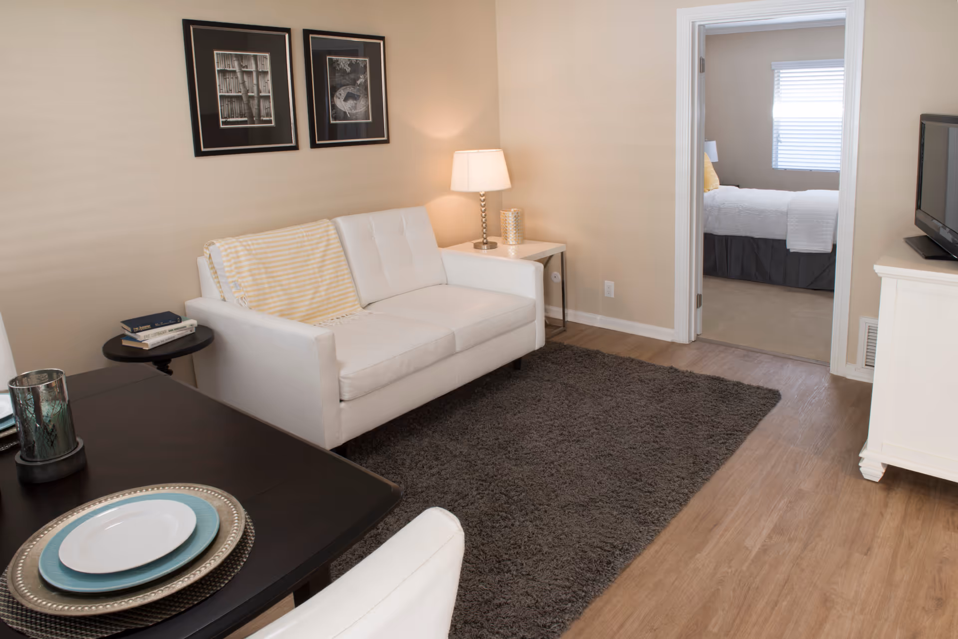 A cozy living room with a white couch draped with a yellow striped blanket, a side table with a lamp and decorative item, two framed black and white pictures on the beige wall, a dark rug on wooden flooring, and a dining table set with plates in the foreground. Through an open doorway, a bedroom with a bed, white bedding, and a window with blinds is visible.