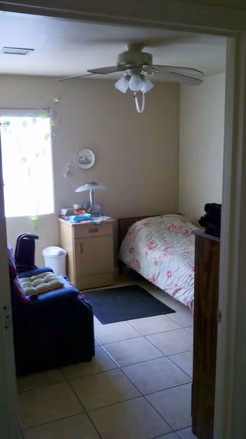 Small bedroom in a senior living unit with bed, dresser, nightstand, chair, and ceiling fan.