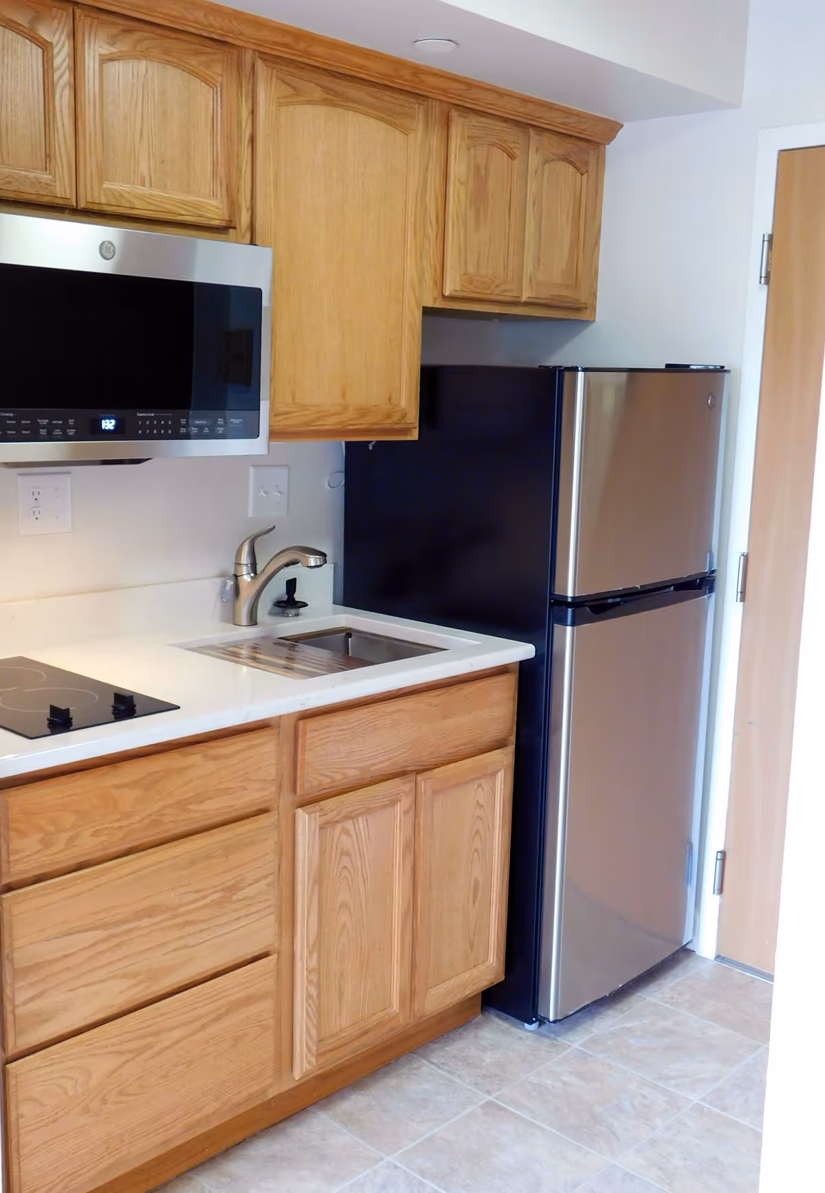 Small kitchenette with oak cabinets, a microwave, sink, electric cooktop and a stainless steel refrigerator.