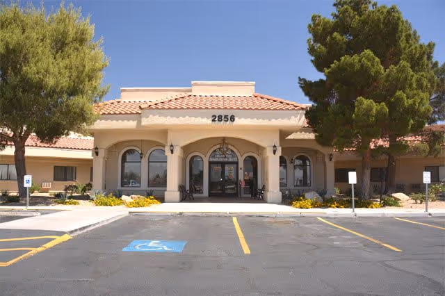 Front exterior view of College Park Rehabilitation Center building with a tiled roof, arched entrance, and several windows. There are trees and landscaping around the entrance, and a parking lot with marked spaces including a handicapped parking spot in front.