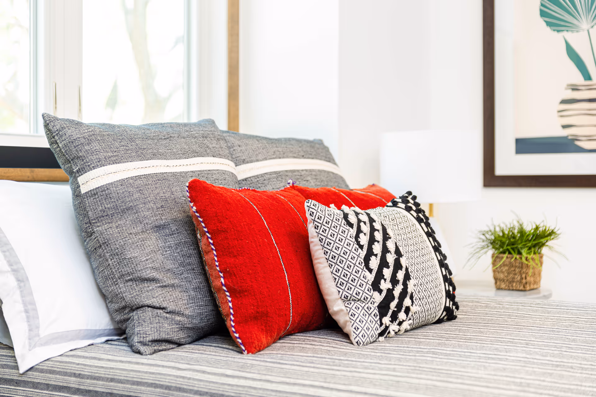 Close-up of a bed with multiple decorative pillows including two large gray pillows with white stripes, two red pillows, and one black and white patterned pillow. The bed has a striped gray bedspread. In the background, there is a white lamp, a small green plant in a woven pot, and a framed artwork on the wall.