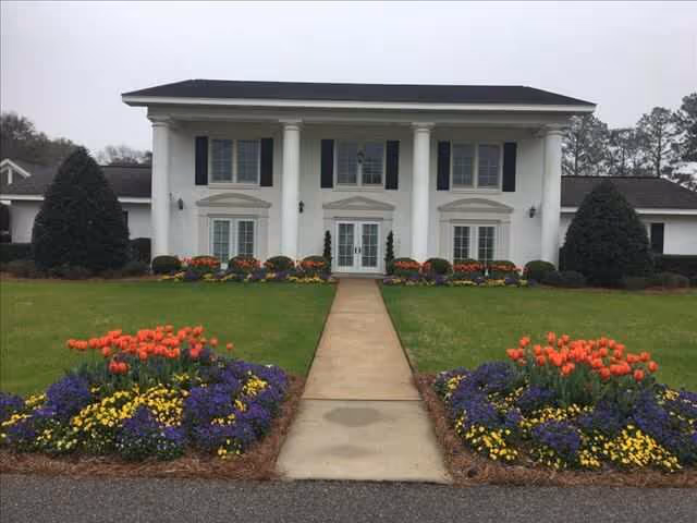 A two-story white building with classical columns, a central entrance, paved walkway, and landscaped flower beds in front.