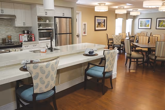 Open communal dining and kitchen area with a long island, upholstered chairs, dining tables, and framed artwork on the walls.