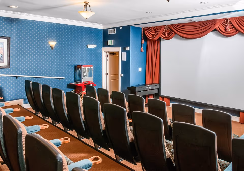 Small screening room with rows of theater seats facing a large projection screen framed by red curtains and a popcorn machine near the entrance.