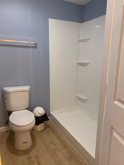 A bathroom with a white toilet, a small trash can with a black liner, a towel rack on the wall, and a white shower stall with built-in shelves. The walls are painted light blue and the floor has a wood-like finish.