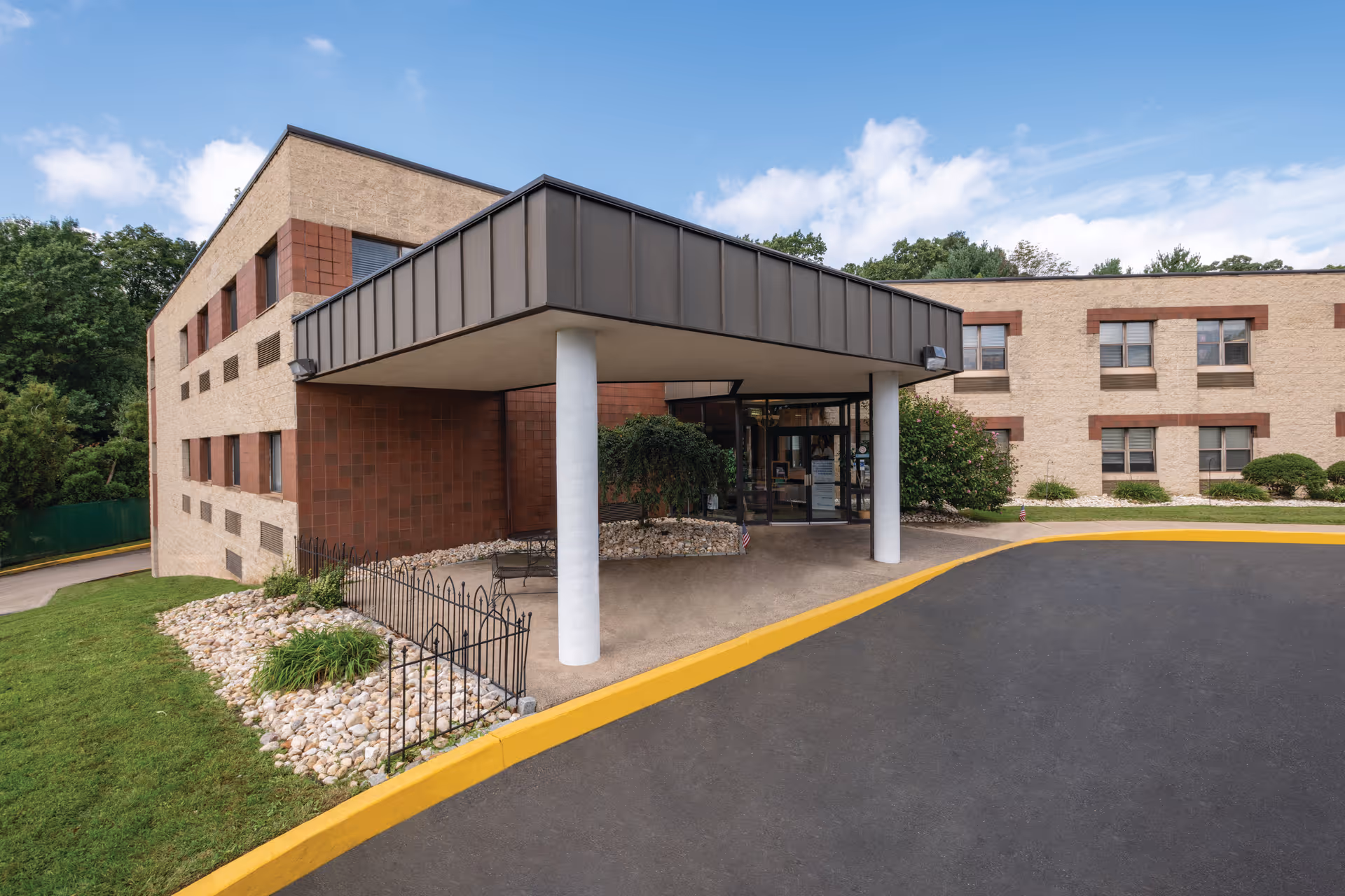 Exterior front view of Dresher Hill Health & Rehabilitation Center showing a two-story building with beige and brown brick walls, a covered entrance supported by two white columns, a paved driveway with a yellow curb, and landscaped areas with grass, bushes, and rocks under a partly cloudy sky.