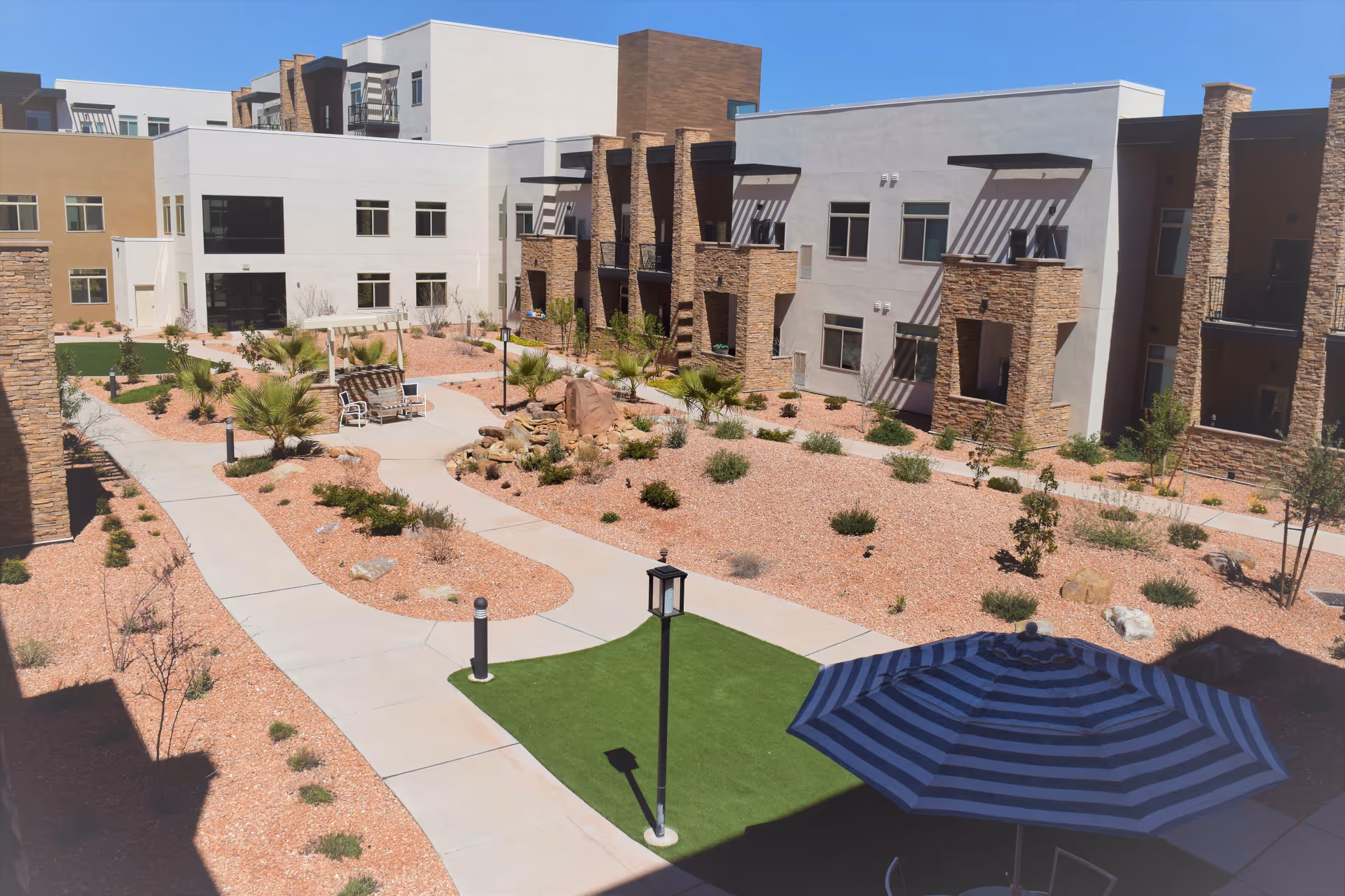 Outdoor courtyard area of a senior living facility with paved walkways, desert landscaping with small bushes and palm trees, a seating area with benches, and a large blue and black striped umbrella providing shade. The surrounding buildings have a modern design with stone and stucco facades and balconies.