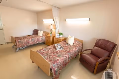 A room with two single beds each covered with patterned bedspreads and white pillows. Between the beds is a wooden nightstand with a lamp, a plant, and a framed photo. There is a brown recliner chair next to one of the beds and a white curtain hanging from the ceiling between the beds. The walls are plain and light-colored, and the floor is a light beige color.