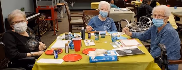 Three elderly women wearing face masks sitting around a table covered with a green tablecloth, engaging in painting and art activities with paintbrushes, paint bottles, paper, and tissue box on the table in a communal indoor setting.