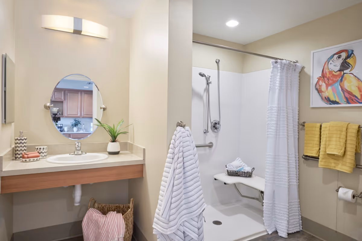 A clean and accessible bathroom featuring a wall-mounted sink with a round mirror above it, a small potted plant, soap dispenser, and a basket with a pink towel underneath. The shower area has a fold-down seat, a handheld showerhead, a white textured shower curtain, and a basket with rolled towels. Yellow towels hang on a rack next to a colorful parrot painting on the wall.