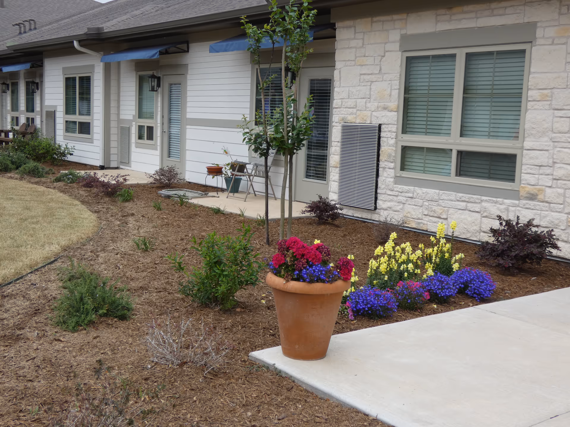 Exterior view of a senior living facility showing a landscaped garden area with a large terracotta pot filled with colorful flowers including red, purple, and yellow blooms. The building has light-colored stone and siding with windows and doors, some with blue awnings above them. There is a concrete walkway in the foreground.