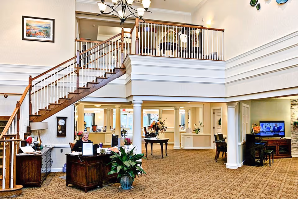 Interior view of a senior living facility lobby with a wooden staircase leading to an upper floor. A receptionist is seated at a wooden desk with a computer, and there are plants and flowers decorating the area. The space has carpeted floors, white columns, and a chandelier hanging from the ceiling. A television is visible in the background on the right side.