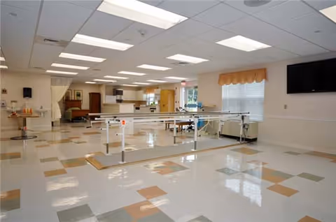Spacious rehabilitation therapy room with parallel bars, tables, and a tiled floor under fluorescent lights.