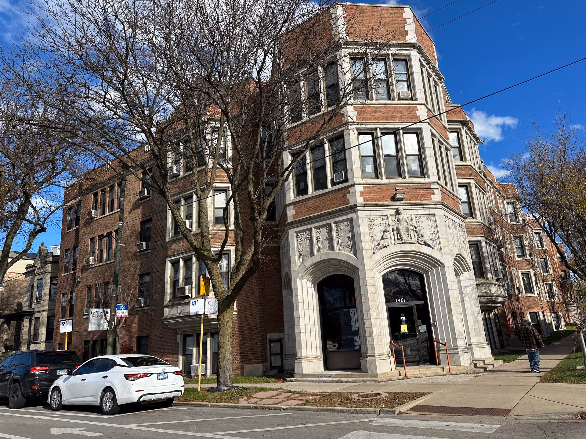 Exterior view of a multi-story brick building with decorative stonework around the entrance. The building has multiple windows with air conditioning units and is situated on a street corner with parked cars and leafless trees. A person is walking on the sidewalk nearby under a partly cloudy blue sky.