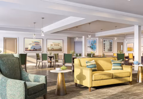 A bright and spacious common area featuring a yellow sofa with two striped pillows, a green upholstered armchair, and several small round tables with decorative items. In the background, there are multiple dining tables with green chairs and framed pictures of vintage vans and scenic views on the walls. The room has a modern design with pendant lighting and wood flooring.