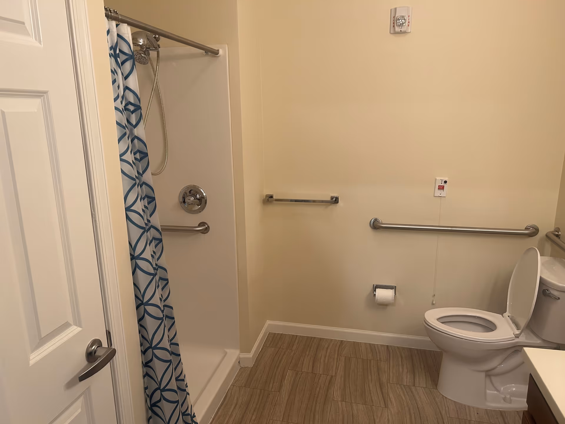 A bathroom with a white toilet, a shower with a blue and white patterned curtain, beige walls, and wood-look tile flooring. There are metal grab bars on the walls near the toilet and inside the shower for accessibility.