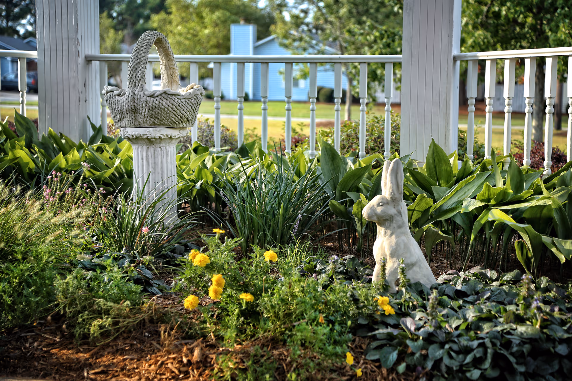 A garden bed with yellow flowers, a white rabbit statue and a decorative basket on a pedestal in front of a white porch railing.