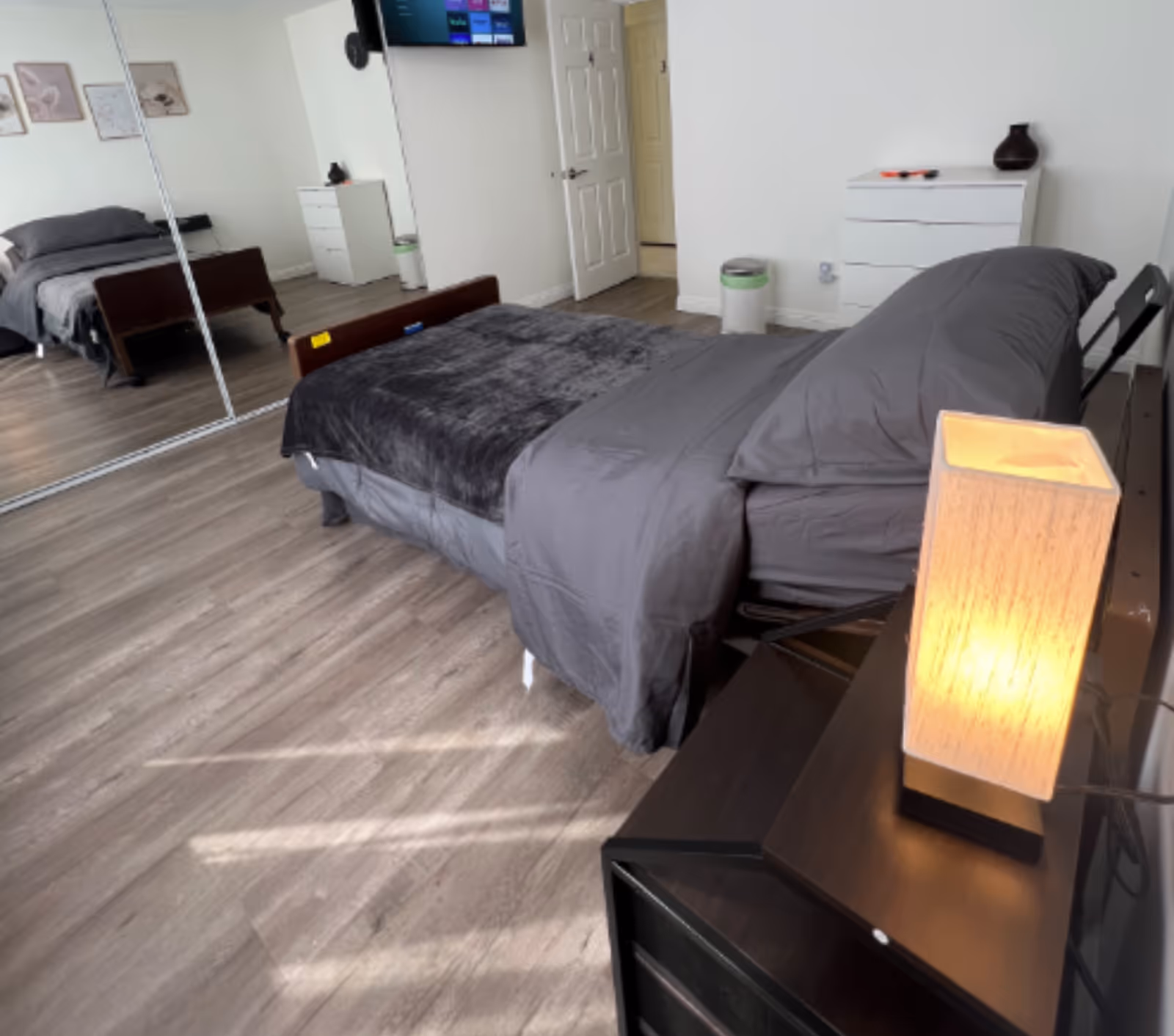Bright bedroom with a made bed, mirrored closet doors, dresser and a lit bedside lamp.