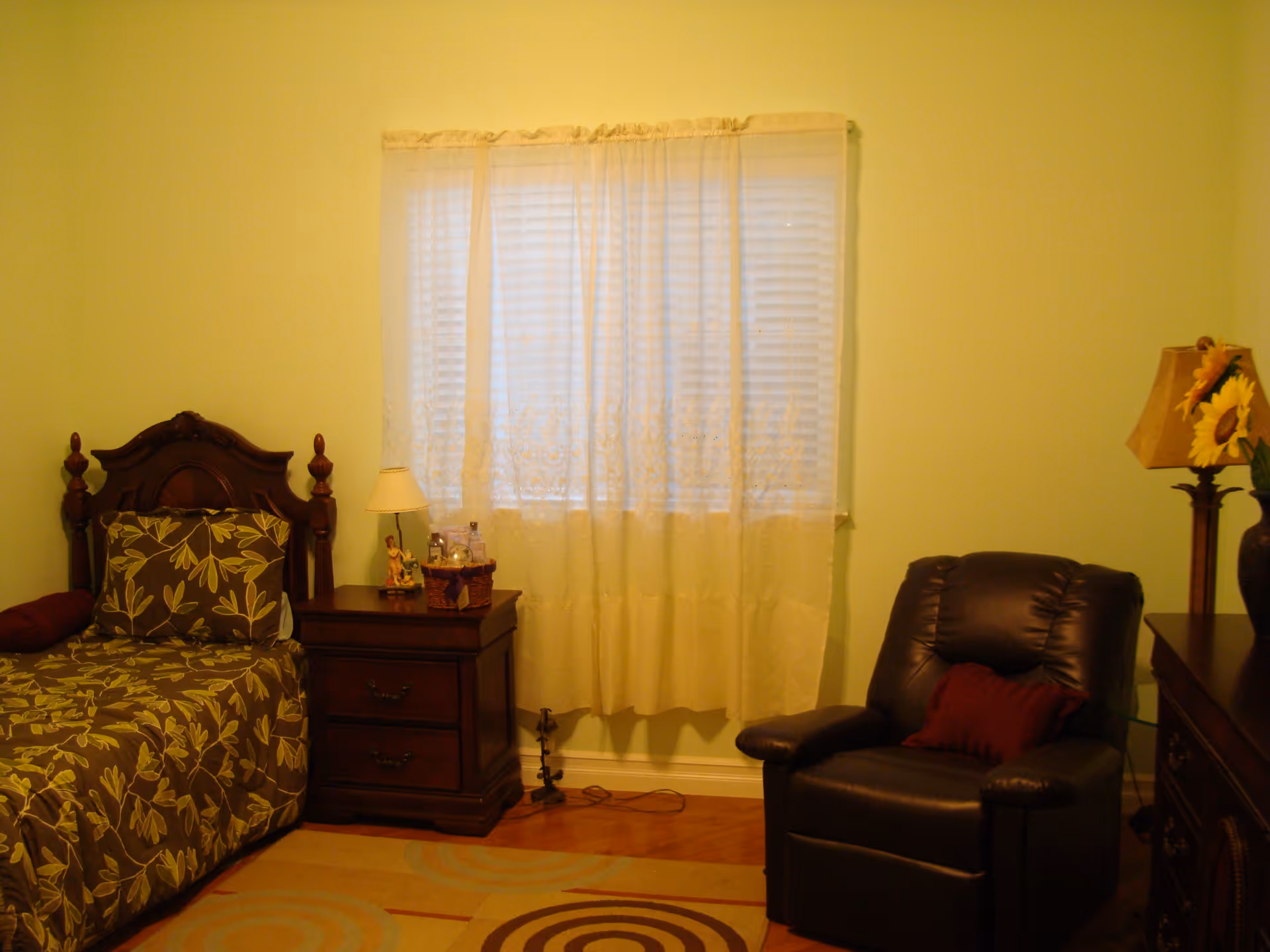 A cozy bedroom with a single bed featuring a floral patterned bedspread and matching pillow. Next to the bed is a wooden nightstand with a small lamp, a figurine, and some personal items. A window with sheer white curtains is centered on the wall behind the nightstand. To the right, there is a dark leather recliner chair with a red cushion. A wooden dresser with a lamp and a vase of sunflowers is partially visible on the right side of the image. The walls are painted light green and the floor has a patterned area rug.
