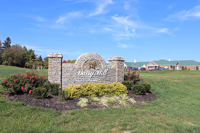 Stone entrance sign reading "Daisy Hill" set in landscaped grass with the senior living building visible in the background under a blue sky.