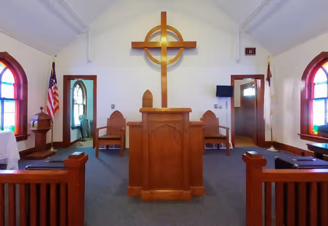 Interior view of a small chapel or worship room with a wooden pulpit in the center, a large wooden cross mounted on the wall behind it, two wooden chairs on either side, stained glass windows on both sides, and flags near the walls.