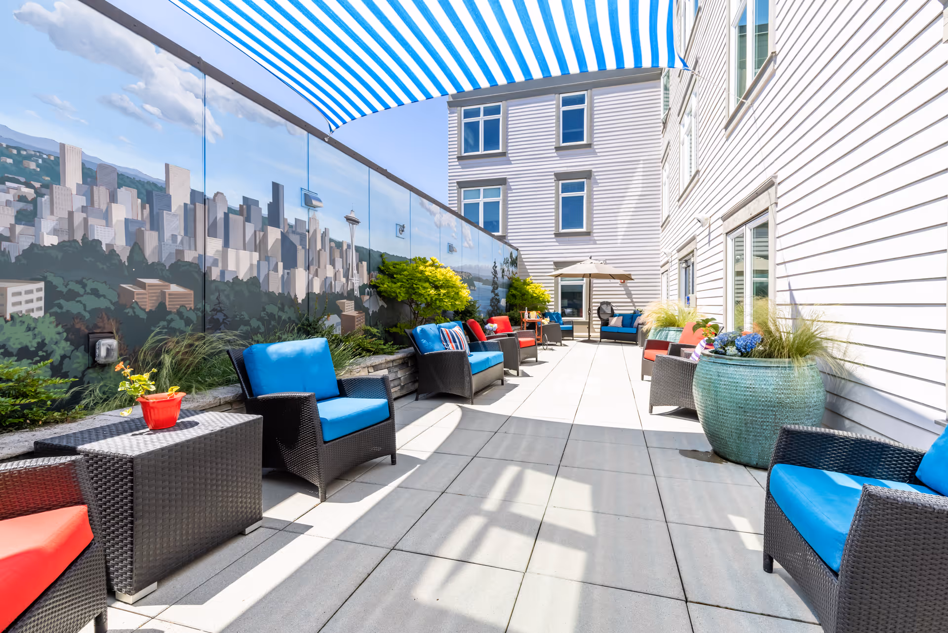Outdoor patio area at Aegis Living Queen Anne Galer with wicker chairs featuring blue and red cushions, small tables, large planters with greenery, and a mural of a city skyline on one wall. A blue and white striped canopy provides partial shade.