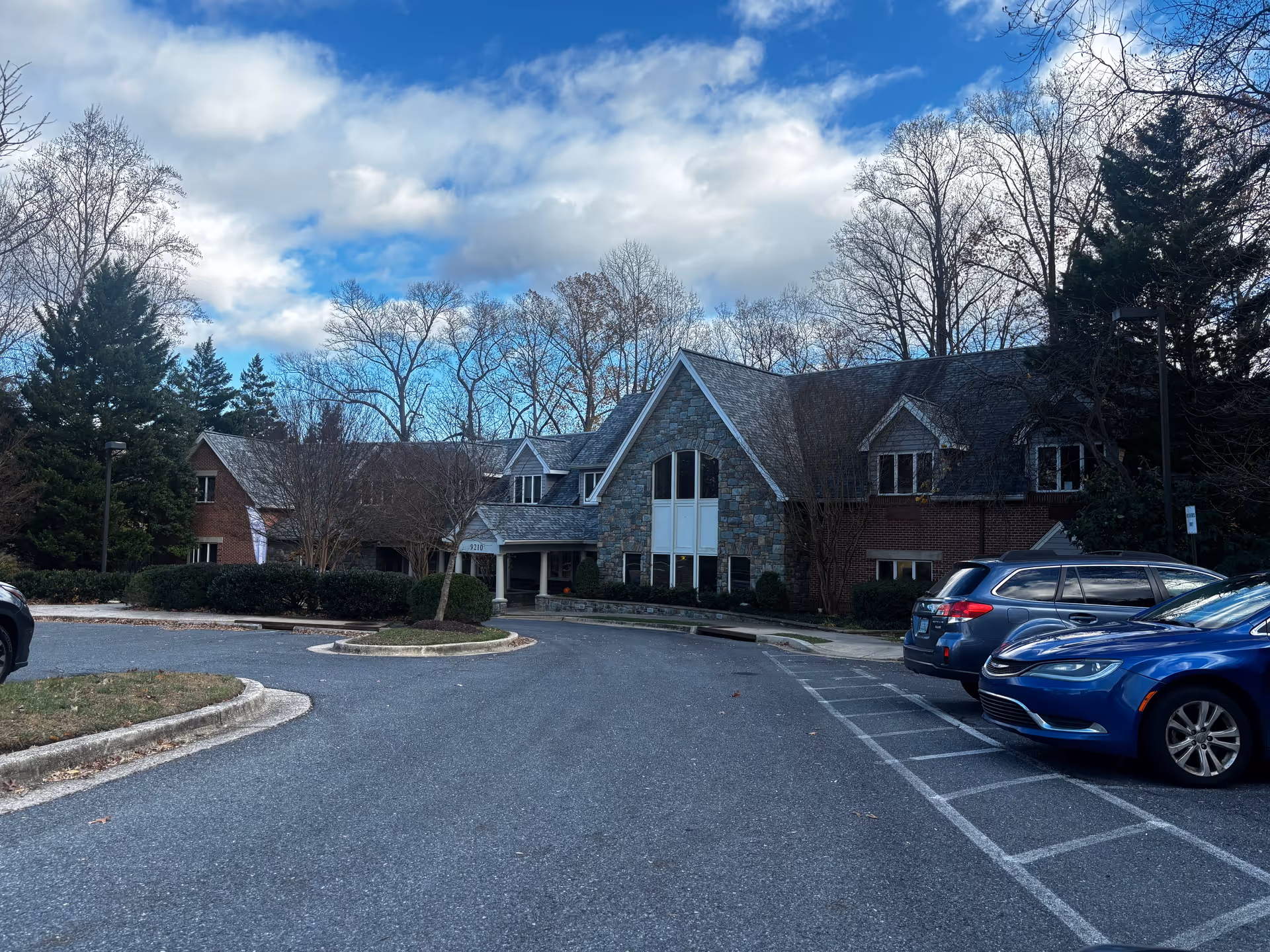 Exterior view of Byron House, a large building with a stone and brick facade, surrounded by leafless trees and parked cars under a partly cloudy blue sky.