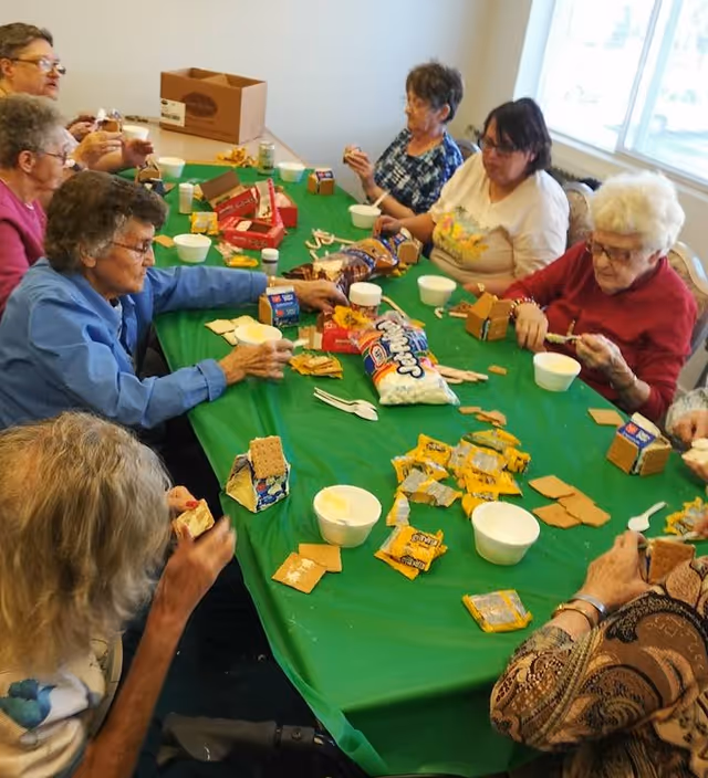 A group of elderly people sitting around a table covered with a green tablecloth, making gingerbread houses using graham crackers, frosting, marshmallows, and candy. The room is well-lit with a window in the background.