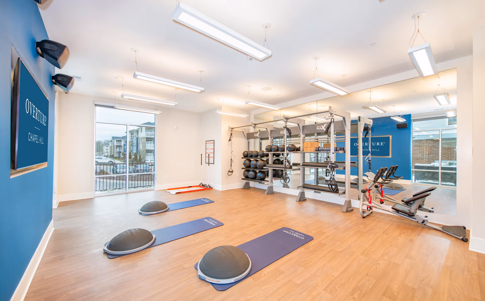Bright fitness room at Overture Chapel Hill with exercise mats, balance trainers, a weight rack, and cardio machines in front of a mirrored wall.