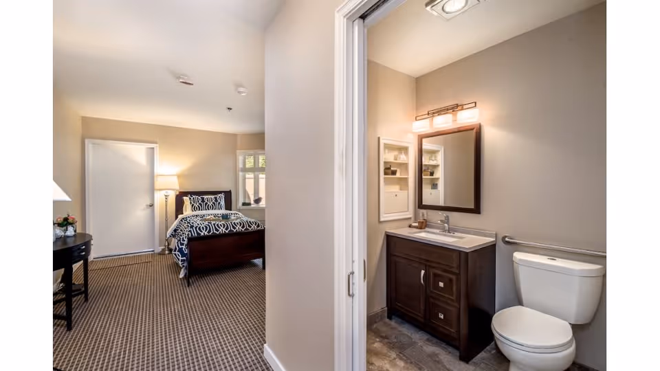 View of a bedroom and an adjacent bathroom in a senior living facility. The bedroom features a bed with patterned bedding, a bedside lamp, a small table with flowers, and a window. The bathroom includes a vanity with a sink, a mirror with lighting above, a toilet with a grab bar, and a small wall cabinet.