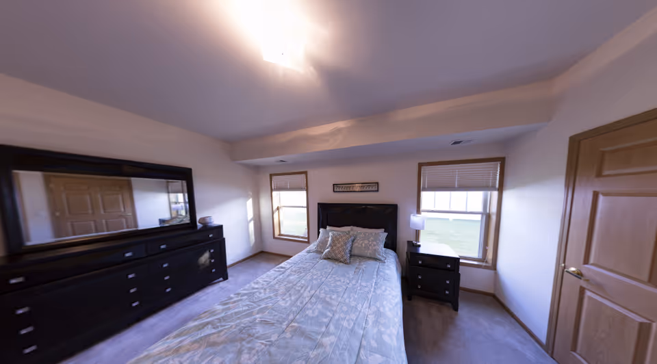 A bedroom with a bed covered in a light blue patterned comforter and two pillows. The room has two windows with blinds, a wooden door, a black dresser with a large mirror, and a black nightstand with a lamp.