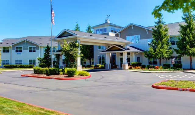 Exterior view of a senior living facility building with a covered entrance, surrounded by landscaped greenery and trees under a clear blue sky. An American flag is raised on a flagpole near the entrance.