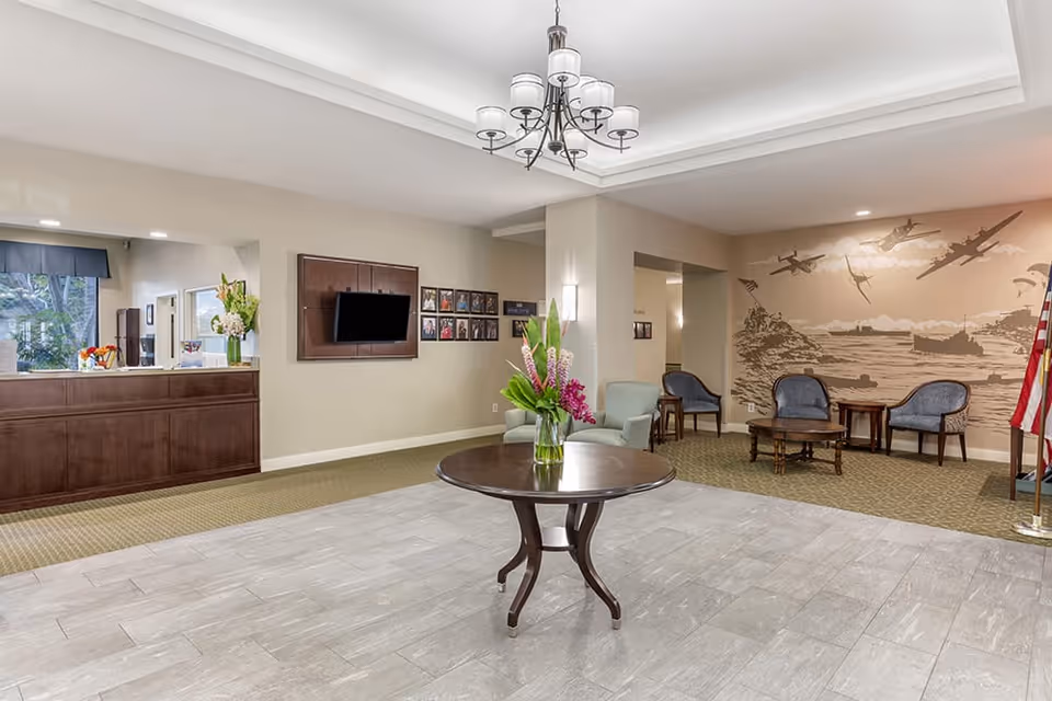 A spacious senior living facility lobby with a round wooden table holding a vase of flowers in the center. To the left is a wooden reception desk with flowers and a window behind it. On the wall, there is a mounted flat-screen TV and several framed photos. To the right, there is a seating area with blue upholstered chairs and wooden tables, and a mural depicting airplanes flying over a coastal scene. An American flag stands in the corner. The ceiling features a chandelier and recessed lighting.