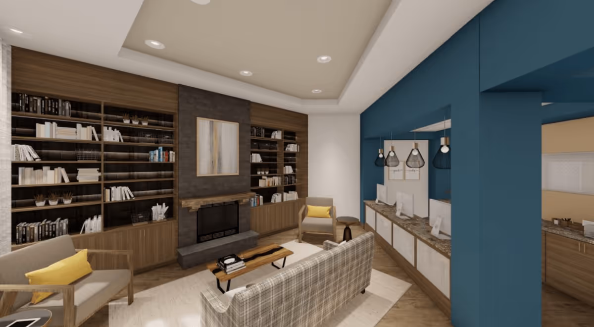 A cozy living room area with a plaid sofa, two armchairs with yellow cushions, a wooden coffee table, and built-in bookshelves flanking a fireplace. On the right side, there is a blue accent wall with a counter featuring three computer workstations and hanging pendant lights above.