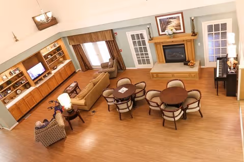 A cozy living room area with wooden flooring featuring a fireplace with a mantel, a piano, two round tables each surrounded by four chairs, a sofa, two armchairs, a TV mounted on a wooden built-in shelf, and large windows with curtains allowing natural light.