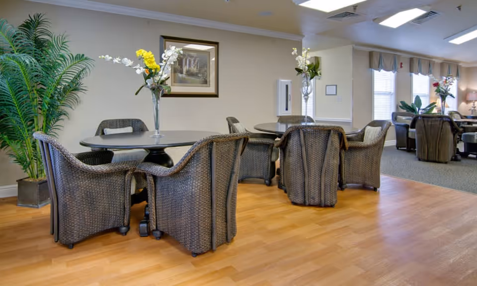 A cozy interior common area with round tables surrounded by wicker chairs, each table decorated with a vase of flowers. The room has light-colored walls, wooden flooring, and large windows with curtains letting in natural light. There are also some potted plants and framed artwork on the walls.