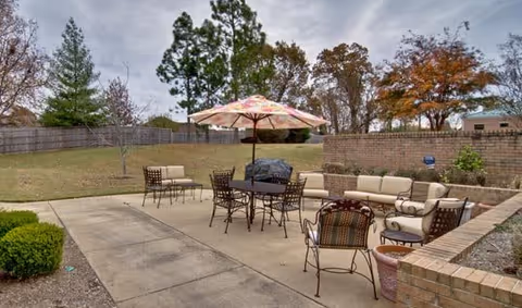 Outdoor patio area with cushioned seating, metal chairs, a table with a colorful umbrella, and a grill. The patio is surrounded by a low brick wall and overlooks a grassy yard with trees and a wooden fence.