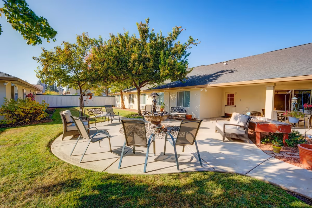Outdoor patio area with a circular concrete seating arrangement featuring metal chairs and cushioned armchairs around a central decorative fountain. The patio is surrounded by green grass, trees, and shrubs, with a single-story building in the background under a clear blue sky.