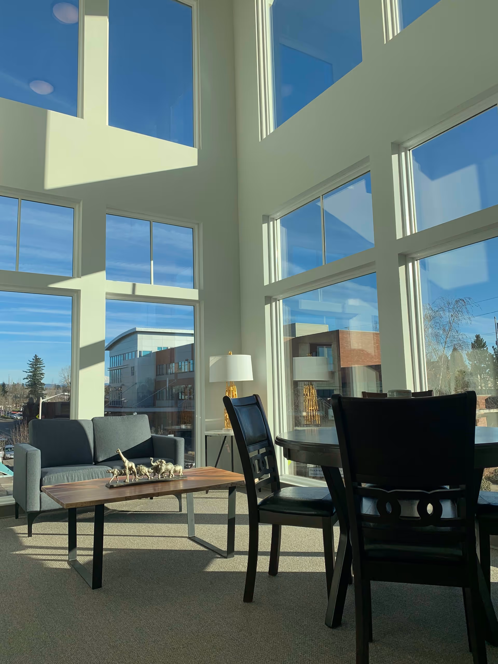 Sunlit lounge with floor-to-ceiling windows, a sofa, coffee table with decorative figurines, dining chairs and a lamp.