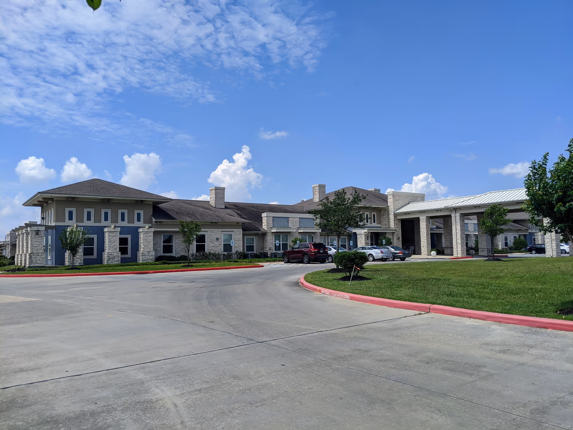 Exterior view of Orchard Park at Victory Lakes Assisted & Senior Living facility showing a large building with stone and beige siding, multiple windows, a covered entrance, several parked cars, green lawn, and a partly cloudy blue sky.