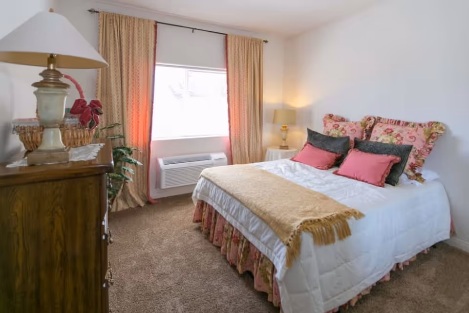 A cozy bedroom with a neatly made bed featuring floral and solid color pillows, a beige throw blanket, and a floral bed skirt. There is a wooden dresser with a lamp and a basket on top, a window with beige curtains, and a small round table with a lamp in the corner. The room has carpeted flooring and white walls.
