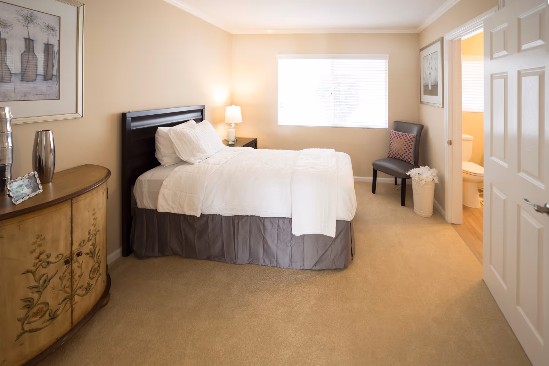 A cozy bedroom with beige walls and carpet, featuring a neatly made bed with white bedding and a dark wooden headboard. To the left is a decorative wooden cabinet with vases and a framed picture above it. A bedside table with a lamp is next to the bed. A gray chair with a colorful pillow and a white vase with flowers are placed near a window with white blinds. An open door reveals a bathroom with a toilet.