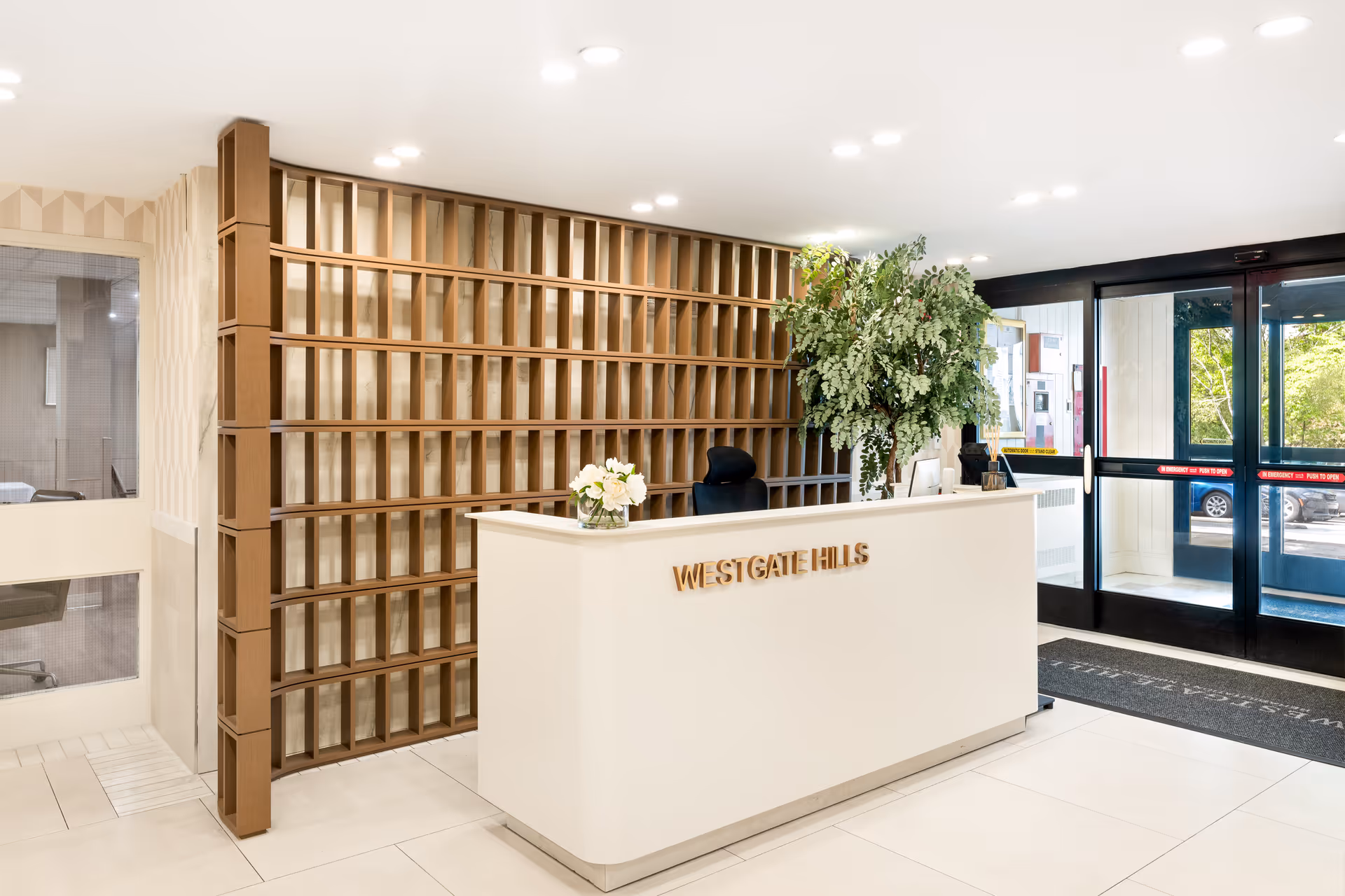 Reception area of Westgate Hills Rehabilitation & Nursing Center featuring a white reception desk with the facility name in gold letters, a large green plant on the desk, a modern wooden shelving unit behind the desk, and glass entrance doors letting in natural light.