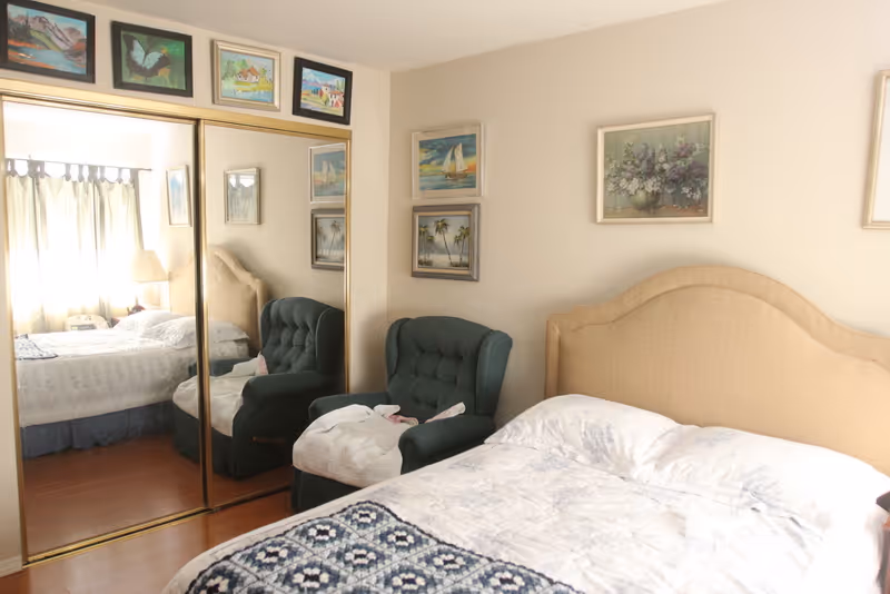 A cozy bedroom with a beige upholstered headboard bed covered with white and blue bedding. Next to the bed is a dark green cushioned armchair with a white blanket draped over it. The wall behind the chair and bed features several framed paintings, including landscapes and floral art. A large mirrored closet door reflects the bed and window with curtains, allowing natural light to brighten the room.