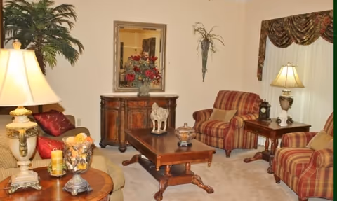 A cozy living room with beige walls and carpet, featuring a beige sofa with red and gold cushions, two red and gold striped armchairs, a wooden coffee table with decorative items, a wooden side table with a lamp, a wooden cabinet with a floral arrangement and a horse figurine, a wall mirror, a wall-mounted plant holder with greenery, and a large potted plant in the corner.