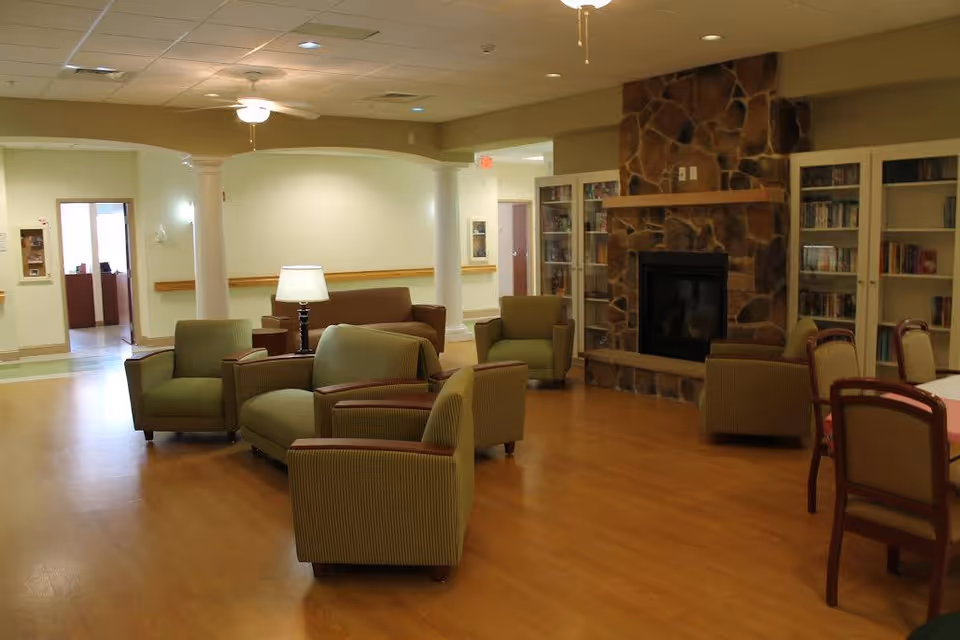 A spacious nursing home common room with green armchairs, sofas, a stone fireplace and bookshelves.