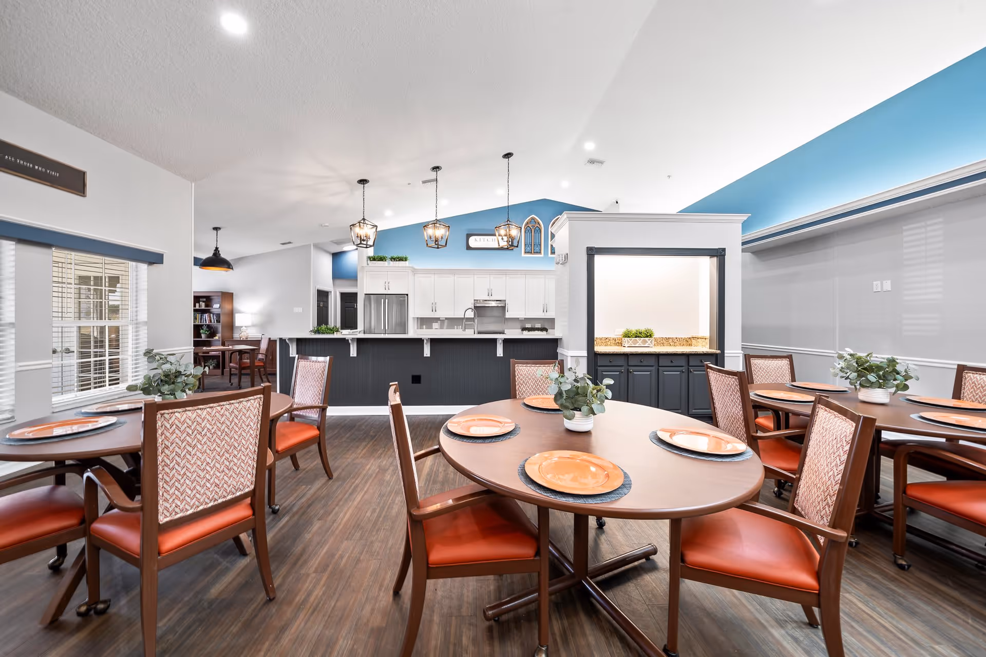 A bright dining room with round wooden tables set with orange plates and small potted plants. The room has wooden flooring, large windows with blinds, and a kitchen area in the background with white cabinets, stainless steel appliances, and pendant lights hanging from the ceiling.